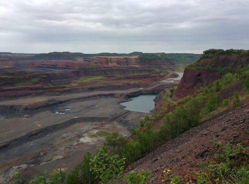 A vast, rugged landscape of a mining site with steep cliffs, a small lake, and overcast skies.