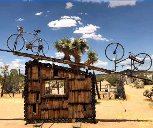 A wooden structure with a window, topped by two bicycles, set against a blue sky with scattered clouds and desert vegetation.
