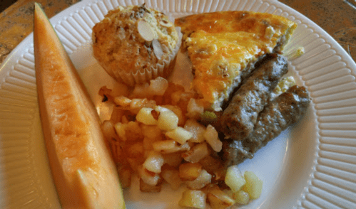 A plate with a slice of omelet, sausage links, fruit salad, a muffin, and a wedge of cantaloupe.