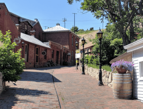 A charming brick pathway lined with historic buildings and lampposts, surrounded by greenery on a sunny day.