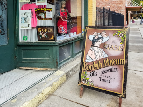A sign for the Oasis Bordello Museum, advertising gifts, souvenirs, and tours, outside a storefront.