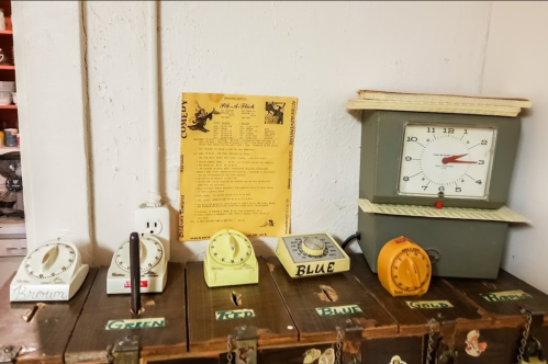 A collection of vintage timers and a clock on a wooden surface, with a yellowed paper listing names and colors.