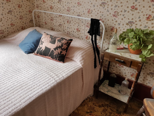 Cozy bedroom with a floral wallpaper, a bed with a pink blanket, decorative pillows, and a small side table with plants.