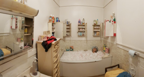 A small bathroom with a bathtub, shelves filled with toiletries, and a mirror reflecting the room's layout.