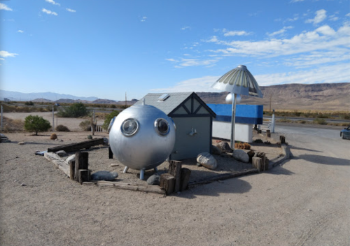 A quirky metallic structure resembling a round spaceship next to a small house in a desert landscape.