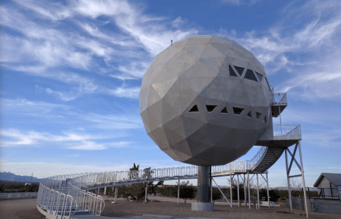 A large, geometric, spherical structure on a raised platform with a spiral ramp, set against a blue sky.