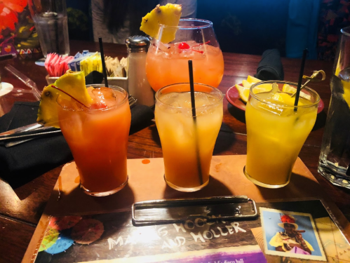 Three colorful cocktails with garnishes, set on a wooden table with a menu and condiments in the background.