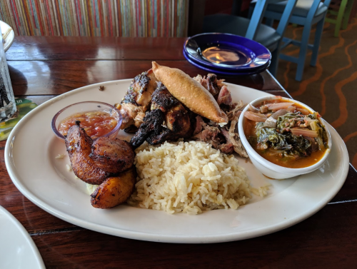 A plate of grilled chicken, rice, fried plantains, and a side of beans with greens, served with salsa.