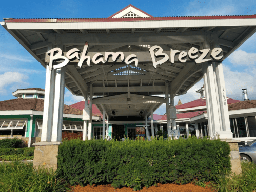 Exterior view of a Bahama Breeze restaurant with a large sign, surrounded by greenery and a blue sky.