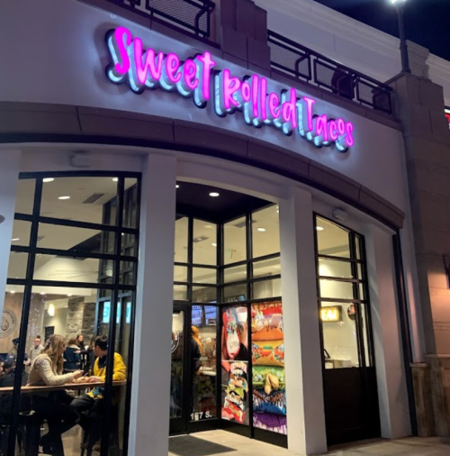 Exterior of "Sweet Rolled Tacos" restaurant at night, featuring colorful signage and a bustling interior.
