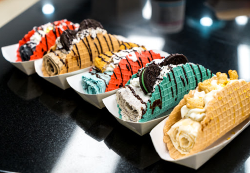 Four colorful waffle tacos filled with rolled ice cream, topped with whipped cream, cookies, and drizzled chocolate.