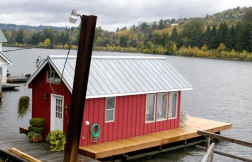 A small red house on a dock by a river, surrounded by trees and hills under a cloudy sky.