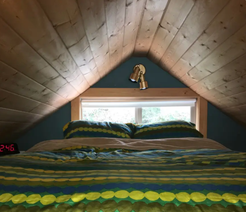 Cozy attic bedroom with a sloped wooden ceiling, colorful bedding, and a window with a warm light fixture above.