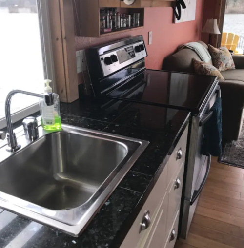 A modern kitchen with a stainless steel sink, black countertop, stove, and a cozy seating area by a window.