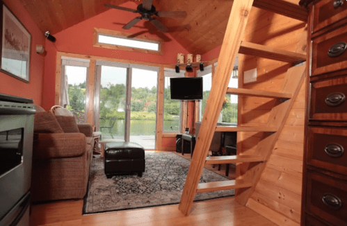 Cozy interior of a cabin with wooden stairs, a sofa, TV, and large windows overlooking a scenic view.
