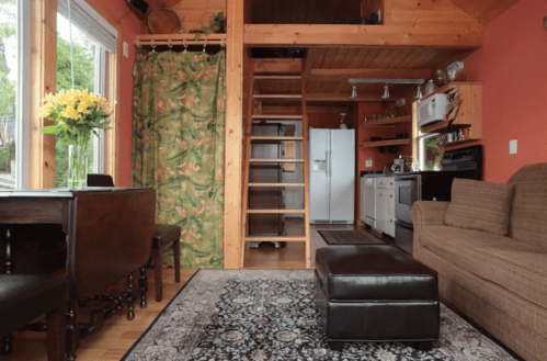 Cozy interior of a small cabin featuring a kitchen, dining area, and a staircase leading to a loft.