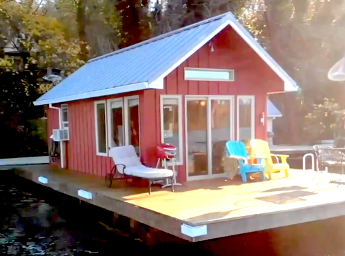 A small red cabin on a dock, featuring large windows, a deck with colorful chairs, and surrounded by trees.