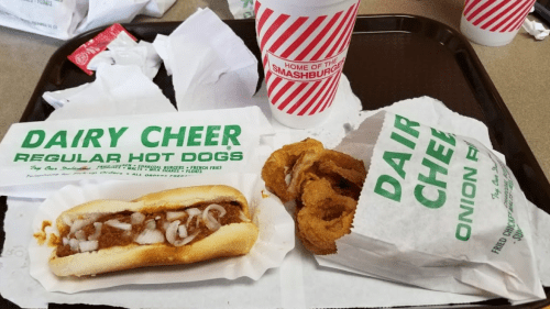 A tray with a hot dog topped with onions, onion rings in a bag, and a drink from Dairy Cheer.
