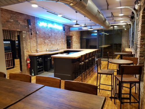 A modern bar interior with wooden tables, high stools, and a brick wall, featuring a neon sign and large windows.