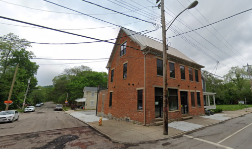 A red brick building at a street corner, surrounded by trees and power lines, with parked cars nearby.