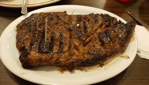 A large, grilled steak on a white plate, garnished with a toothpick, resting on a wooden table.