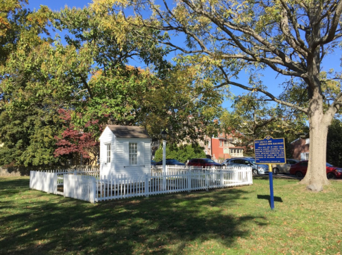 A small white building surrounded by a white picket fence, with trees and a historical marker nearby on a sunny day.
