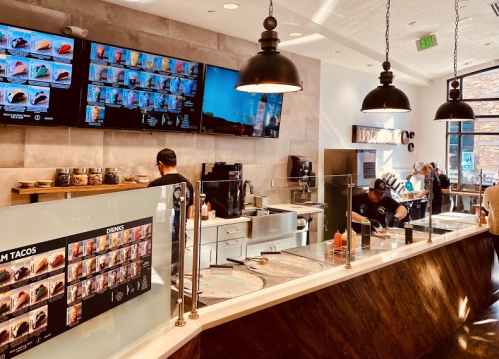 A modern restaurant interior with a counter, menu displays, and staff preparing food in a bright, inviting space.