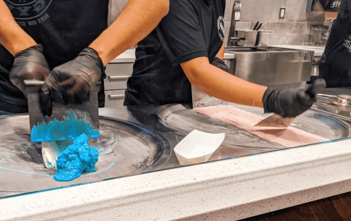 Two people in black gloves prepare colorful ice cream on a cold surface, using metal spatulas.