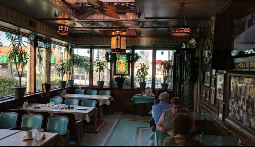 Cozy restaurant interior with green plants, tables, and large windows, creating a warm, inviting atmosphere.