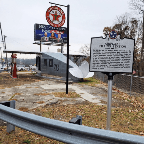 A vintage airplane filling station with a sign, gas pump, and billboard in a rural setting.