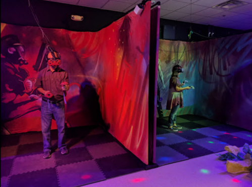 Two people wearing VR headsets stand in a colorful, dimly lit room with vibrant wall art and soft flooring.