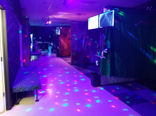 A dimly lit room with colorful lights, benches, and screens, creating a vibrant atmosphere for activities.