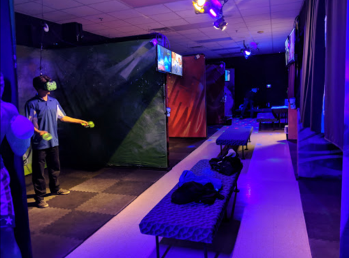 A dimly lit VR gaming room with a player using a headset, benches, and screens displaying game visuals.