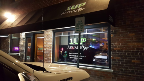 Exterior view of 1UP Arcade Bar at night, featuring a sign and illuminated windows showcasing arcade games and cocktails.