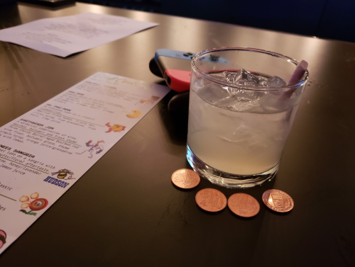 A cocktail with ice sits on a table next to a menu and three coins, with colorful game controllers in the background.