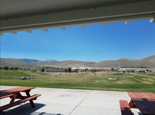 View of a grassy landscape with mountains in the distance, framed by a covered patio and picnic tables in the foreground.