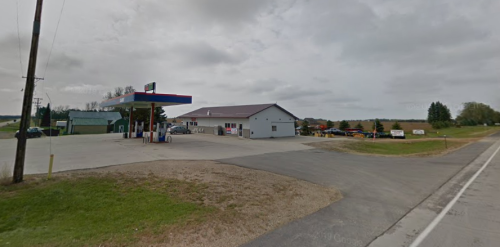 A gas station and convenience store along a rural road, with cloudy skies and grassy areas nearby.
