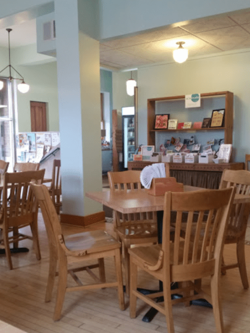A cozy café interior with wooden tables and chairs, featuring a bookshelf filled with books and magazines.