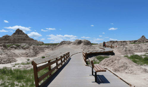 A wooden walkway winds through a rugged landscape of eroded hills under a bright blue sky with scattered clouds.