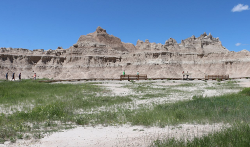 Visitors walk along a wooden path in a grassy area, with rugged rock formations and a blue sky in the background.