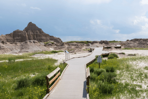 A wooden boardwalk winds through grassy terrain, leading to rocky formations under a cloudy sky.