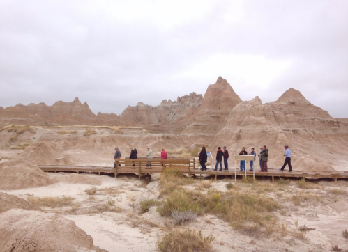 A group of people walking on a wooden boardwalk in a rugged, sandy landscape with unique rock formations.