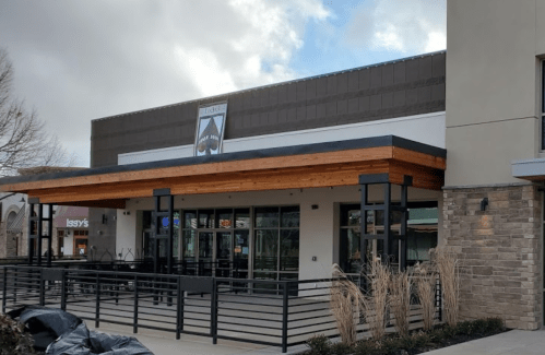 Exterior of a modern café with a wooden overhang, large windows, and a patio area, set against a cloudy sky.