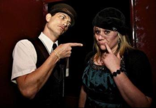 A man in a hat points at a woman in a black dress, who is making a playful, thoughtful expression.