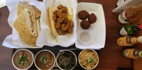 A tray with a sandwich, fried shrimp, and three bowls of sides: beans, collard greens, and rice, with hot sauces on the side.