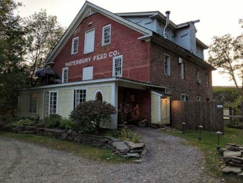 Historic building with a red and yellow exterior, featuring "Waterbury Feed Co." sign and surrounded by greenery.