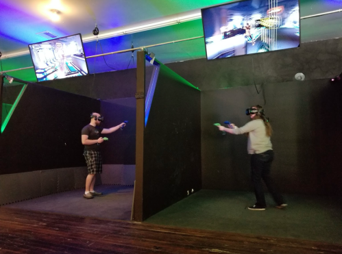 Two people wearing VR headsets play a game in a dark room with screens above them displaying the game.
