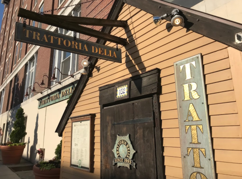 Exterior of Trattoria Delia, featuring a wooden facade, sign, and menu board, with a brick building in the background.