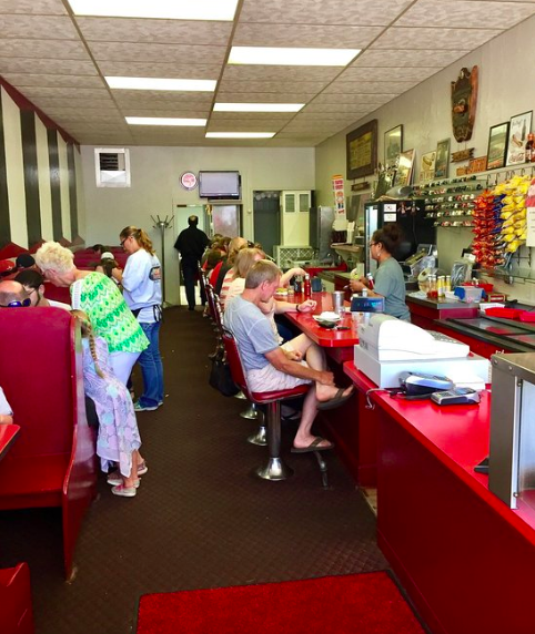 A busy diner interior with red booths and a counter, filled with customers enjoying meals and chatting.