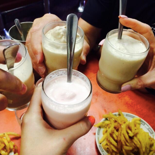 A group of hands holding glasses of creamy drinks, with a plate of fries in the background on a red table.